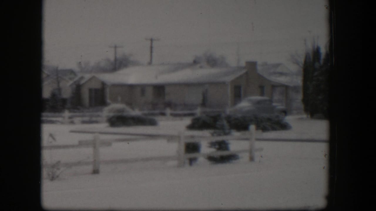 1954 Nevada home-movie view of a snowy residential house, white fence, shrubs, utility poles, and parked mid-century cars under a gray sky.
