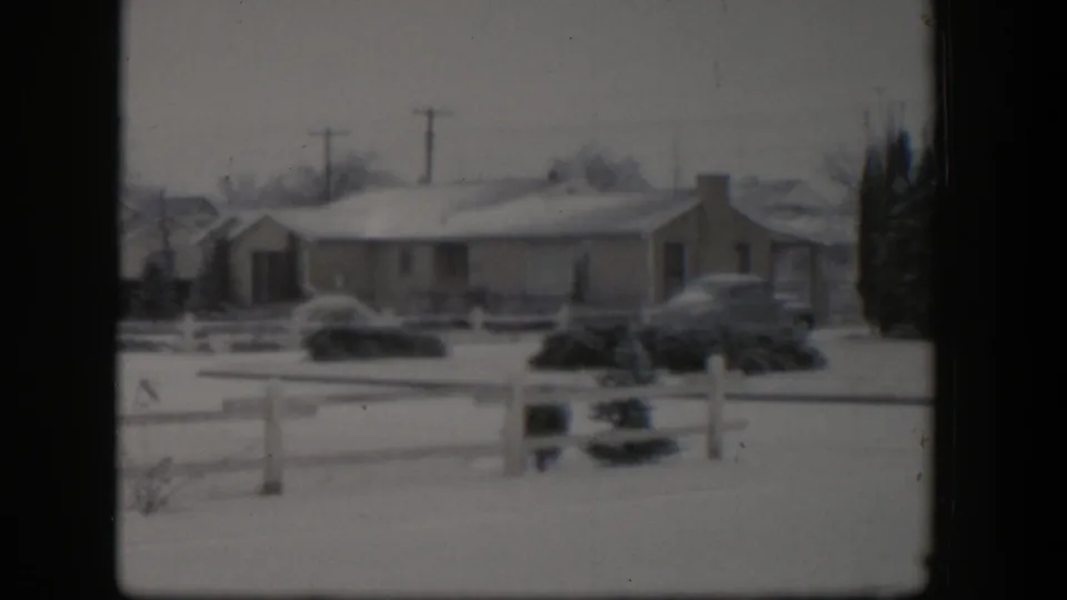 1954 Nevada home-movie view of a snowy residential house, white fence, shrubs, utility poles, and parked mid-century cars under a gray sky.