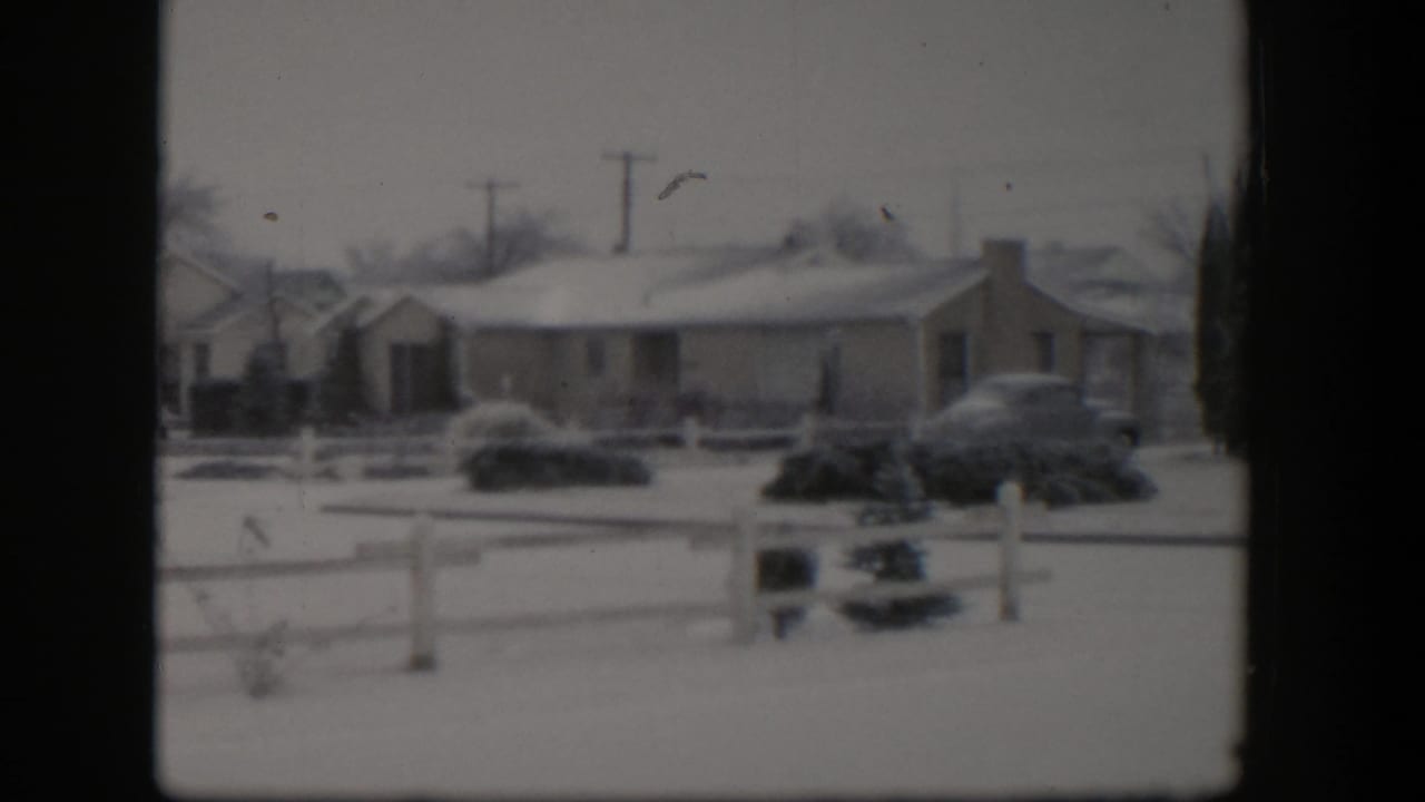 Snowy residential house and shrubs seen across a white fence with parked cars nearby.