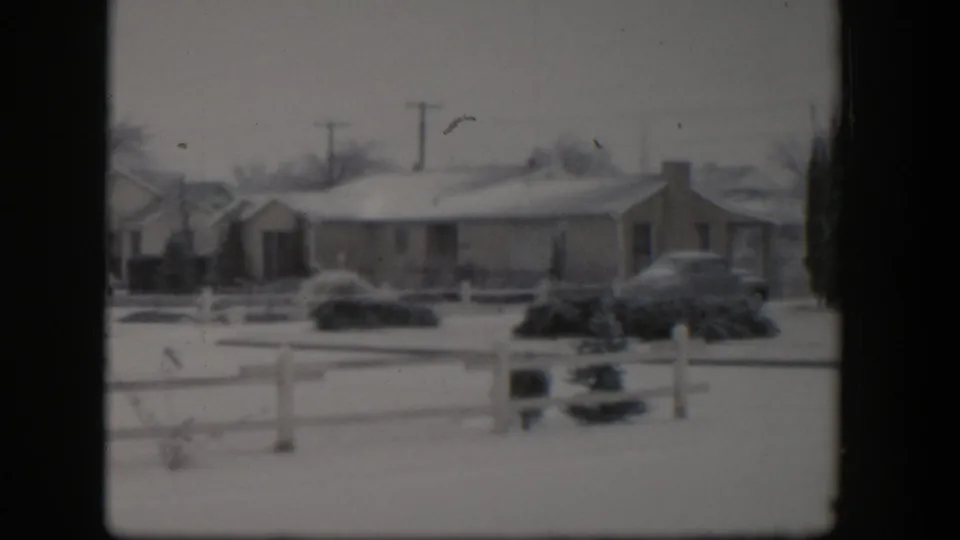 Snow-covered Nevada residential exterior from 1954, showing a low house, white fence, shrubs, parked cars, and utility poles in soft 8mm focus.