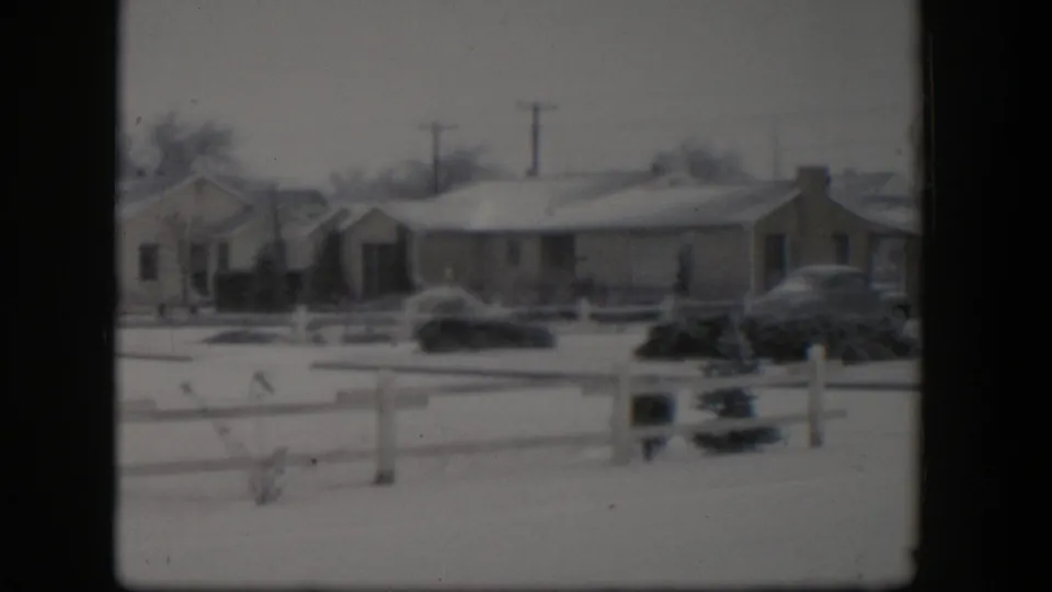 1954 Nevada winter neighborhood view with snow-covered houses, a white fence, low shrubs, a parked car, utility poles, and bare trees.