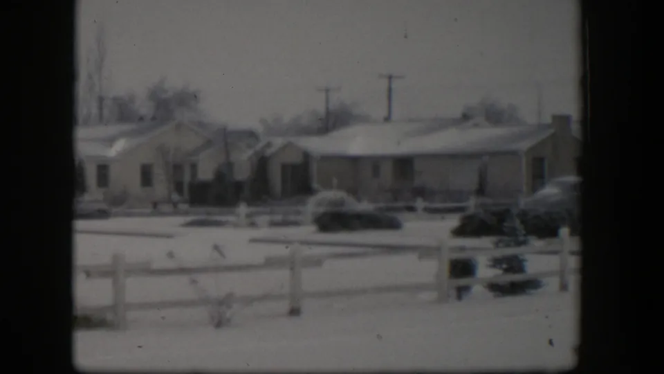Final 1954 Nevada frame showing a snow-covered residential row, white fence, shrubs, parked cars, utility poles, and bare winter trees.