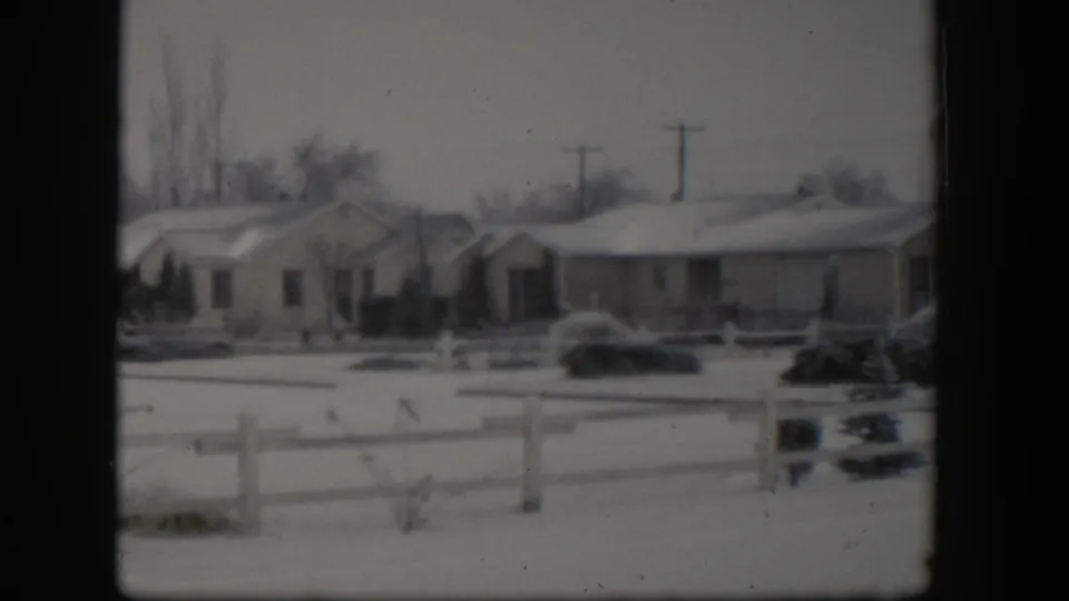 1954 Nevada home-movie frame showing snow-covered single-story houses, a low fence, shrubs, and power lines under a gray sky.