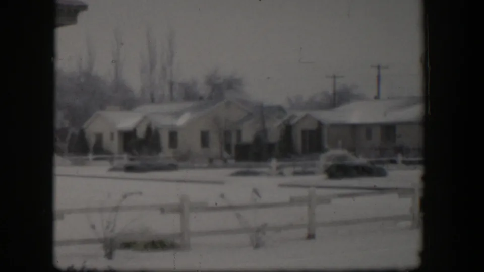 1954 Nevada residential winter scene with snow on roofs and yards, a low wooden fence, shrubs, and utility poles.