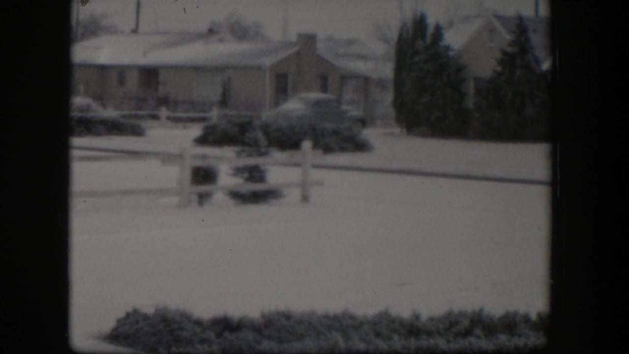 Snow-covered suburban street with a ranch house, parked car, fence post, and evergreens.