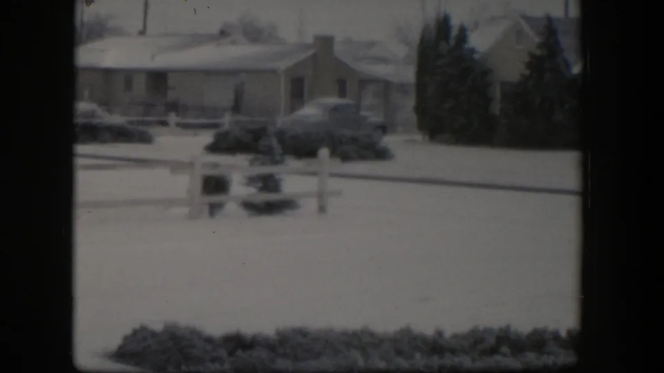 1954 Nevada 8mm frame of a snow-covered suburban street with a ranch-style house, parked car, low fence post, and dark evergreens.