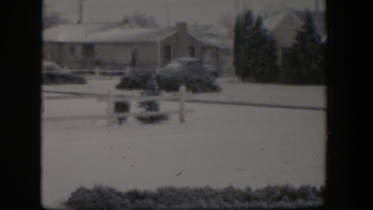 Snowy residential street with ranch house, parked car, shrubs, and dark evergreens.