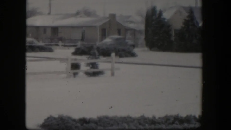 1954 Nevada home-movie frame showing a snow-covered ranch house frontage, a parked mid-century car, shrubs, and evergreen trees.