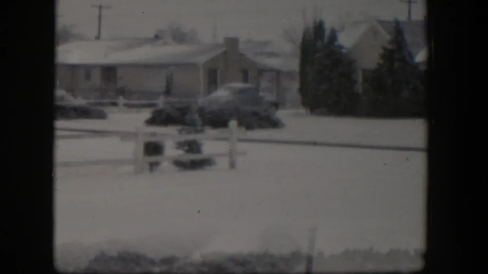 Soft 8mm view of a Nevada suburban ranch house in snow, with a parked car, fence post, shrubs, and dark trees in the yard.