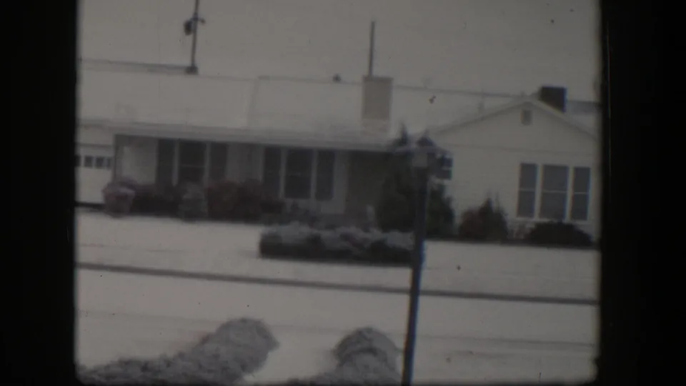 1954 Nevada frame showing another snow-covered ranch-style home with front windows, a chimney, shrubs, street edge, and utility pole.