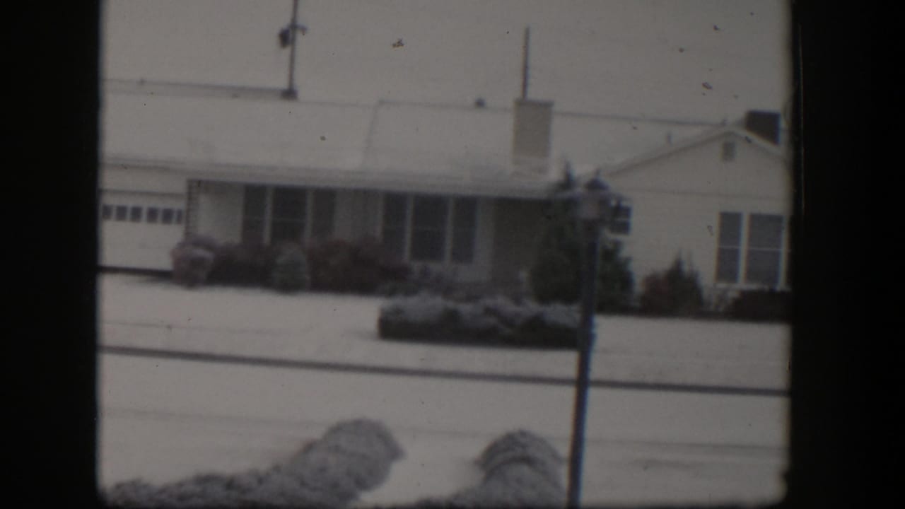 Snow-covered single-story house with garage, shrubs, lamppost, and power lines.