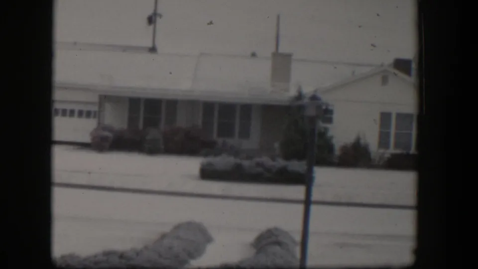 1954 Nevada home-movie frame of a snowy bungalow exterior with garage, front windows, shrubs, and a lamppost.
