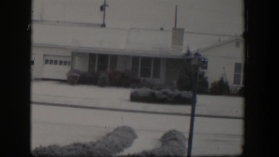 1954 Nevada frame showing a snow-covered residential exterior, attached garage, shrubs, lamppost, and overhead lines.