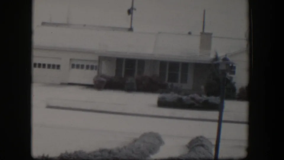 1954 Nevada home-movie frame with a closer view of the snow-covered bungalow facade, garage, windows, and yard shrubs.