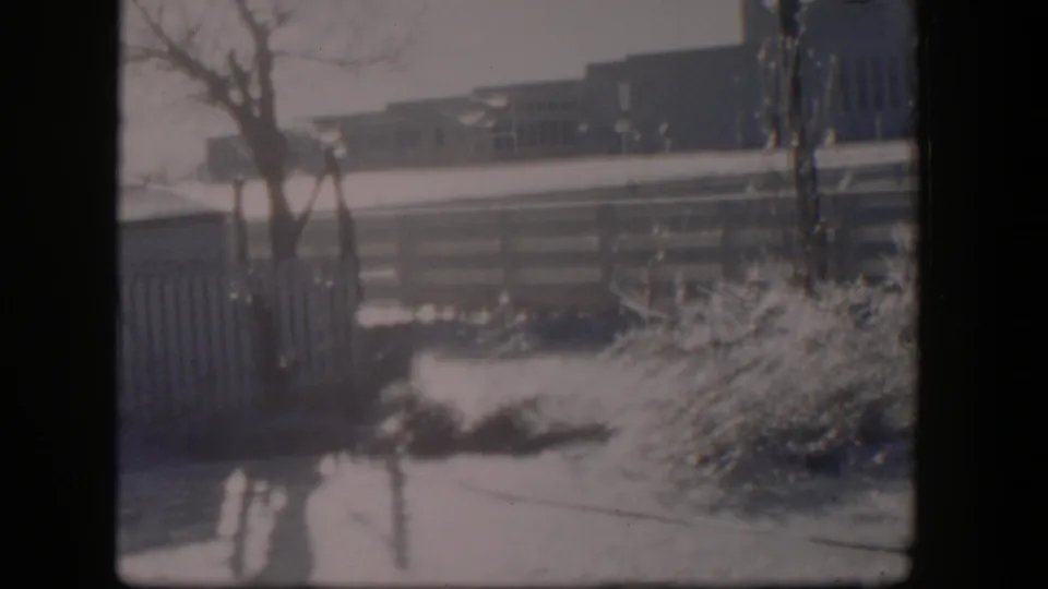 1954 Nevada 8mm frame showing snow on grass and shrubs near a wooden fence, with bare trees and low buildings beyond.