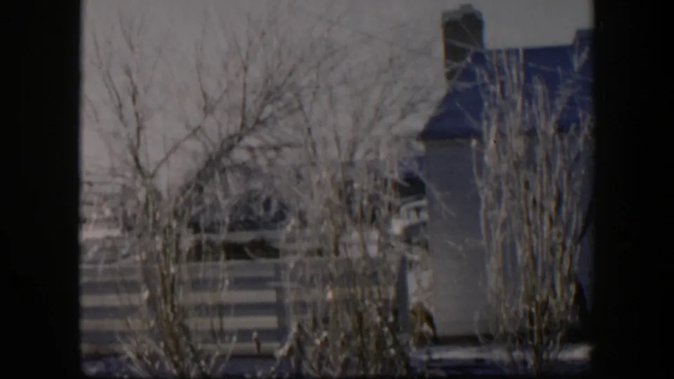 1954 Nevada 8mm frame of a snow-dusted yard with leafless trees, shrubs, a striped fence, and a nearby house exterior.