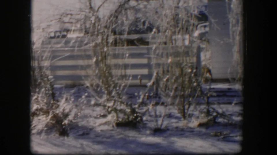 1954 Nevada winter home-movie frame showing snowy ground, bare plantings, a striped fence, a utility pole, and part of a house exterior.