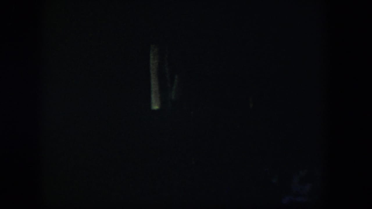 Extremely dim 1954 Nevada home-movie frame with a shadowed room and faint pale vertical forms near the left and center.