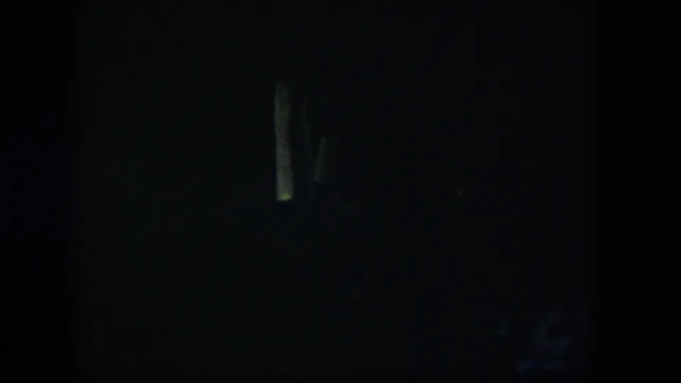 Extremely dim 1954 Nevada home-movie frame with a shadowed room and faint pale vertical forms near the left and center.