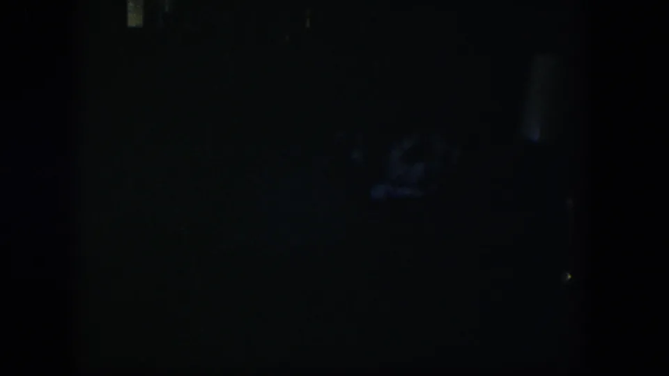 Underexposed 1954 Nevada home-movie frame with a pale central object, possibly a chair or lamp, and faint blue shapes below.