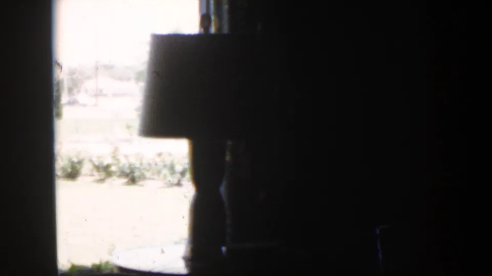 1954 Nevada 8mm frame showing a table lamp in silhouette against a bright window inside a dark home interior.