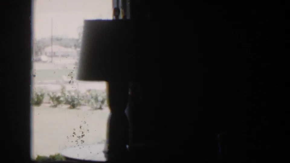 1954 Nevada home-movie frame showing a dark room, lamp silhouette, bright window, lawn, shrubs, and distant houses.