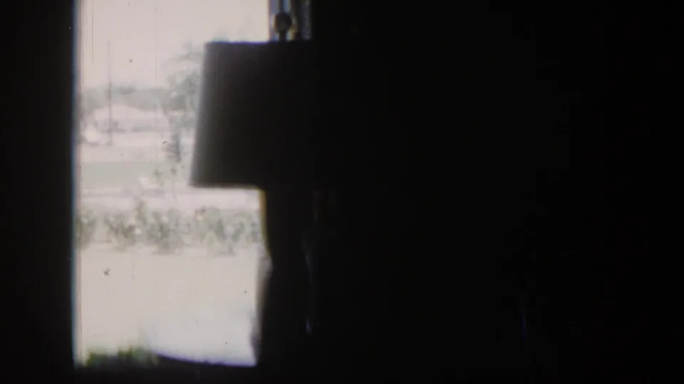 1954 Nevada interior frame with a shaded lamp in silhouette and a washed bright view of lawn and houses outside.