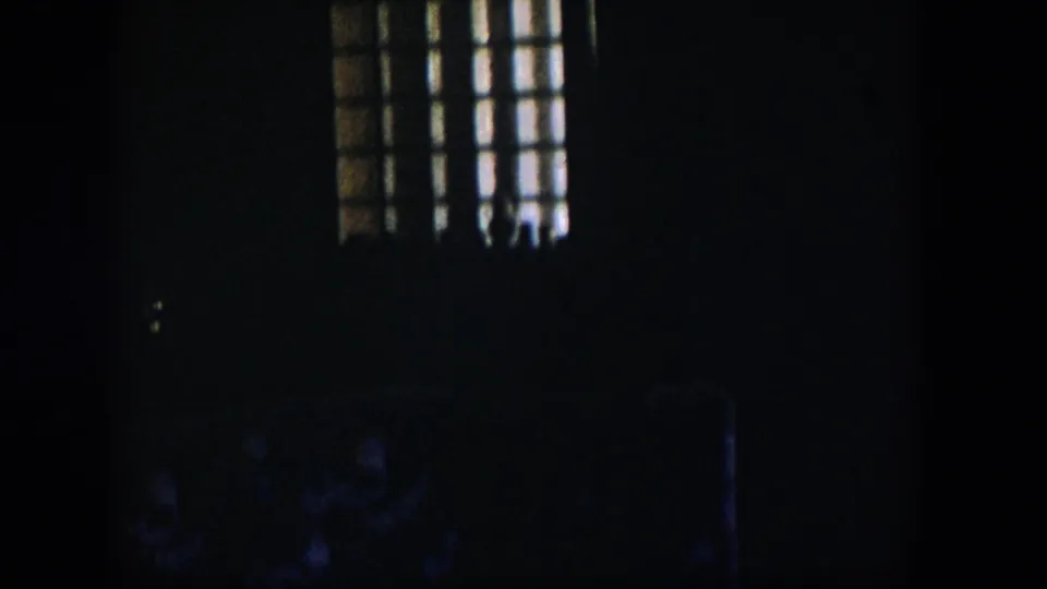 A very dark 1954 Nevada 8mm frame showing light through a barred or gridded window above faintly visible upholstered furniture in a private interior.