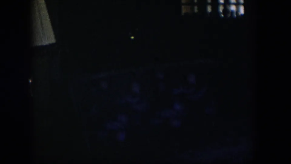 A shadowed 1954 Nevada home-movie frame where a pale lampshade and faint patterned sofa or chair emerge from the dark interior.
