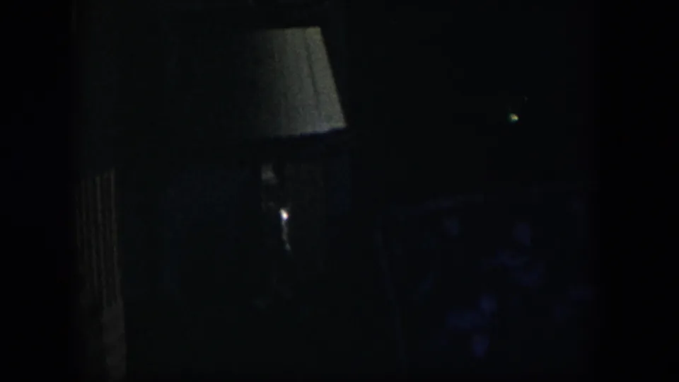 A low-light 1954 Nevada 8mm frame showing a white lampshade, dark vertical surfaces, and indistinct upholstered furniture inside a private room.