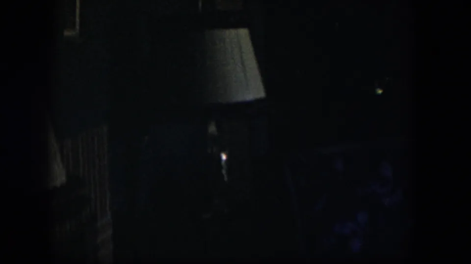 A dark 1954 Nevada home-movie frame with a shaded lamp near center, vertical wall or furniture lines, and faint patterned seating in the lower right.