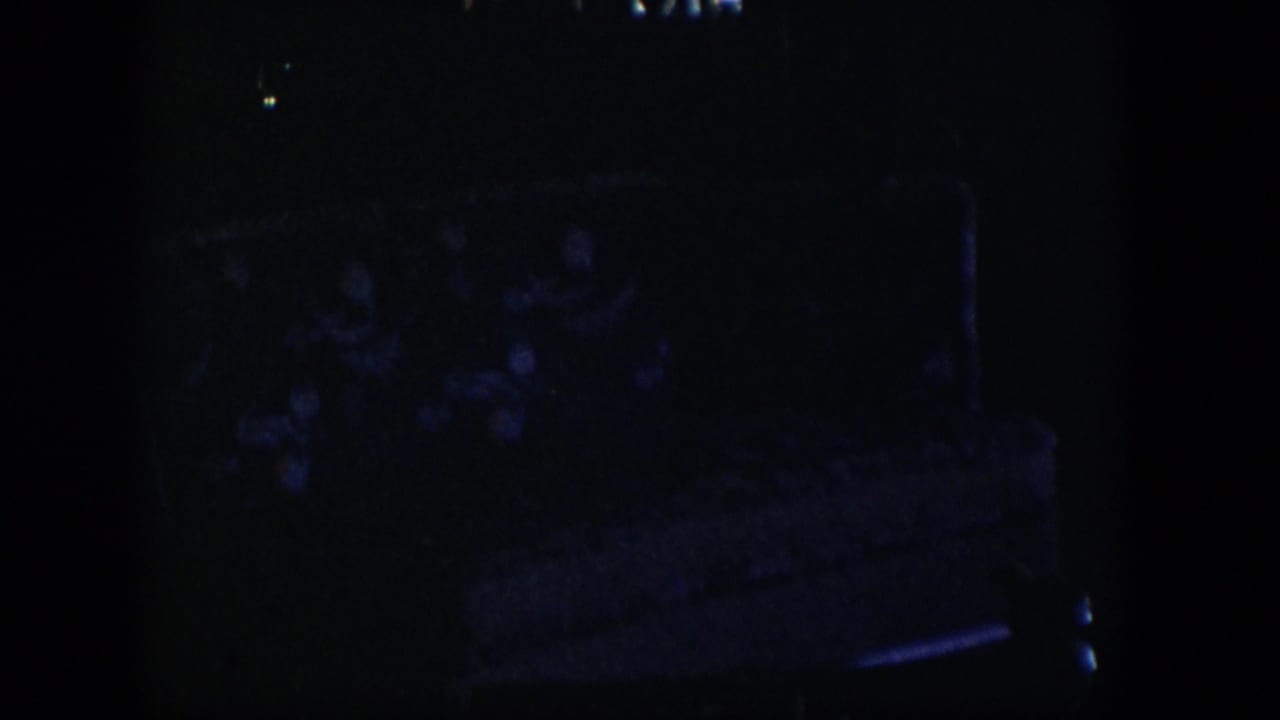 Very dark 1954 Nevada home-movie frame with a faint patterned upholstered bed or chair in deep shadow.