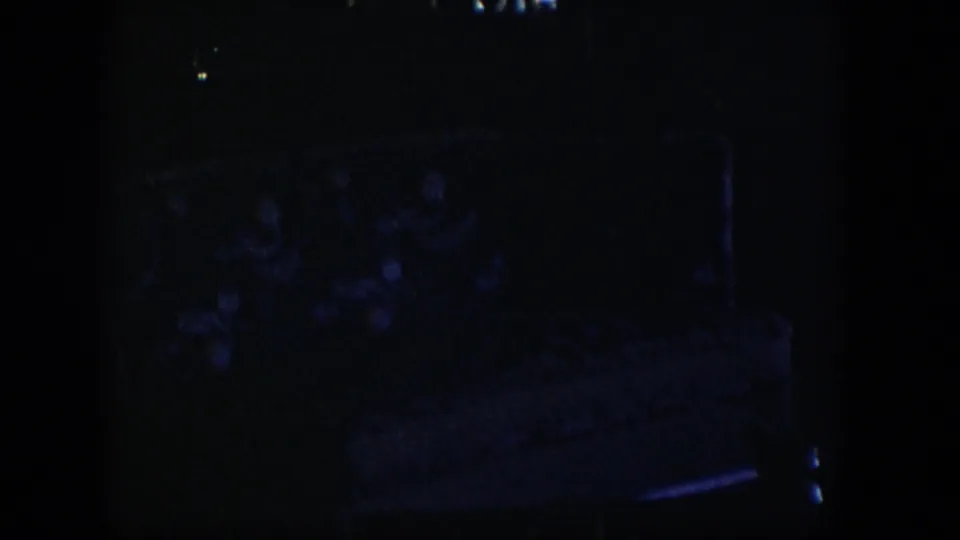 Very dark 1954 Nevada home-movie frame with a faint patterned upholstered bed or chair in deep shadow.