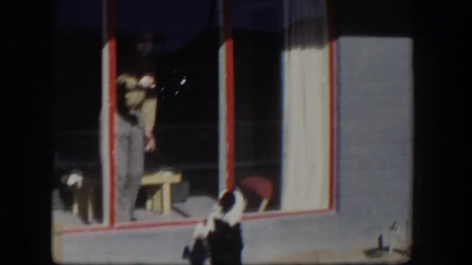 1954 Nevada home-movie frame showing a black and white dog outside a red-trimmed window while a person stands inside near a small table.