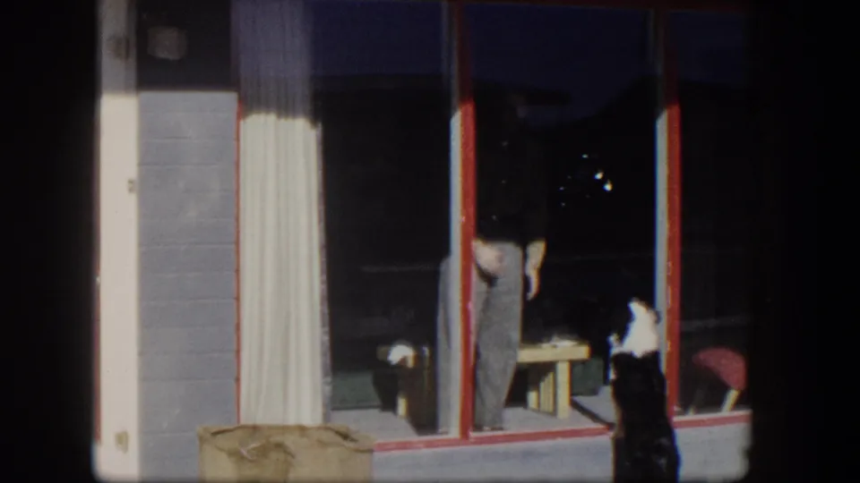 1954 Nevada home-movie frame showing a man inside a glass door and a black-and-white dog outside beside a red door and gray wall.