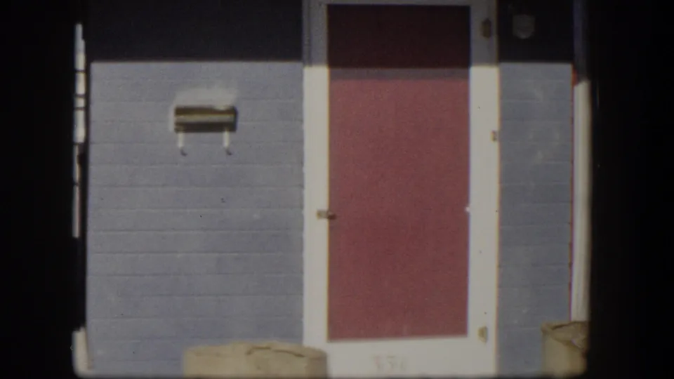 1954 Nevada home exterior showing gray masonry, a red door, a small wall-mounted mail slot, and cream curtain at the edge.