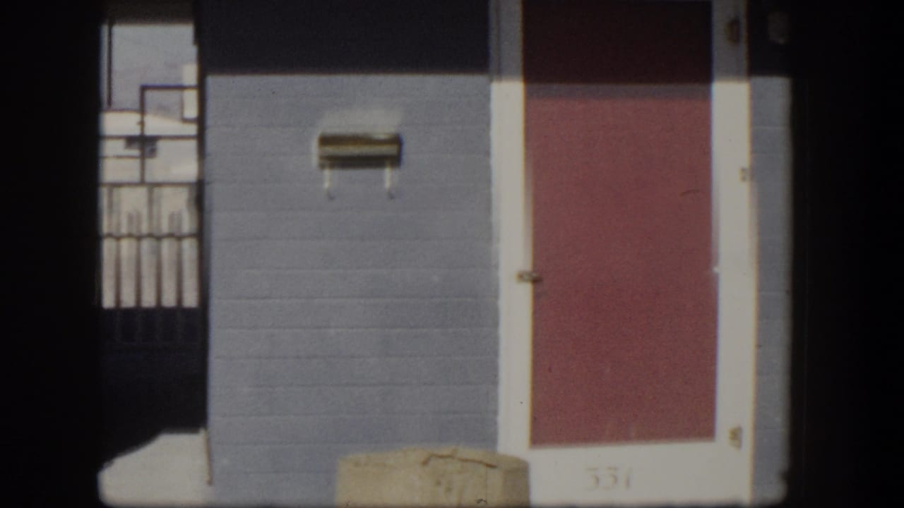 Blue stucco bungalow wall with red door, wall mailbox, and part of a covered carport.