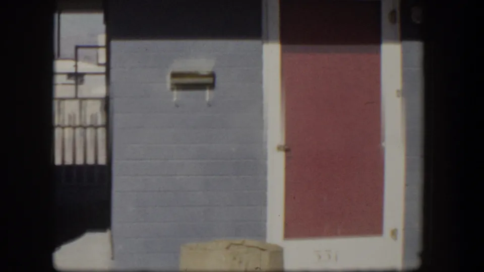 1954 Nevada home-movie frame showing a blue stucco bungalow exterior, red door marked 331, wall mailbox, and carport shadow.