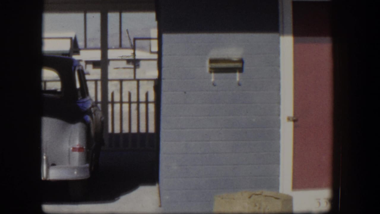 Partial rear view of a silver mid-century sedan parked under a carport beside a stucco wall.