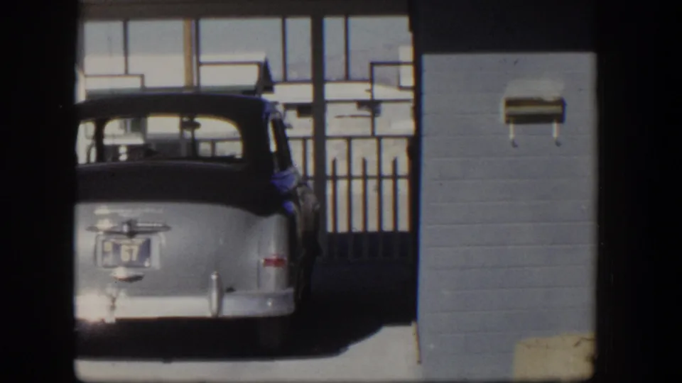 Rear view of a silver 1950s sedan parked beside the blue stucco bungalow exterior in the Nevada home-movie clip.
