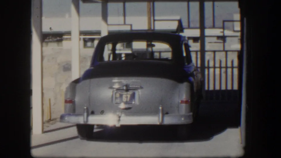 The parked silver sedan is centered under the carport, with blue stucco, fencing, and driveway details visible in the 1954 Nevada footage.