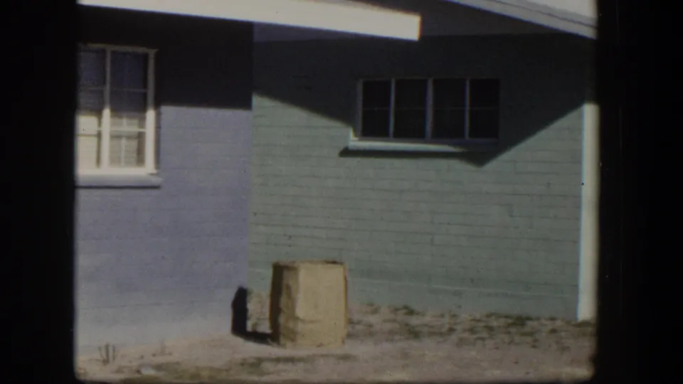 1954 Nevada home exterior showing blue-painted masonry, divided windows, and a bag or sack beside the dusty yard edge.