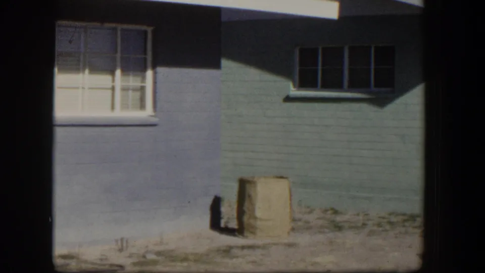 1954 Nevada frame showing a painted residential exterior with window trim, angular roof shadow, and a paper bag on bare ground.