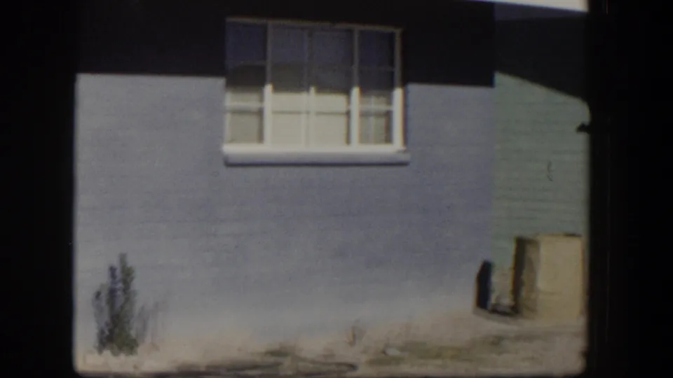 1954 Nevada 8mm view of a blue house wall with a white-trimmed window, closed blinds, sparse weeds, and bare sunlit ground.