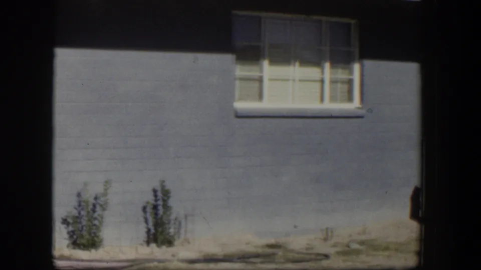 1954 Nevada frame showing the blue house exterior, white window trim, closed blinds, sparse bushes, and sunlit bare ground.