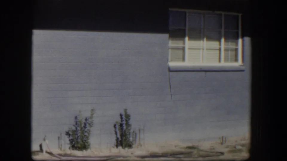 1954 Nevada 8mm frame showing blue-gray house siding, a white-trimmed window, sandy ground and small shrubs near the wall.