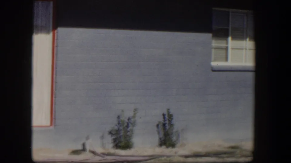 1954 Nevada home-movie frame with a high window, blue-gray siding, pale vertical trim and small shrubs in a dry yard.