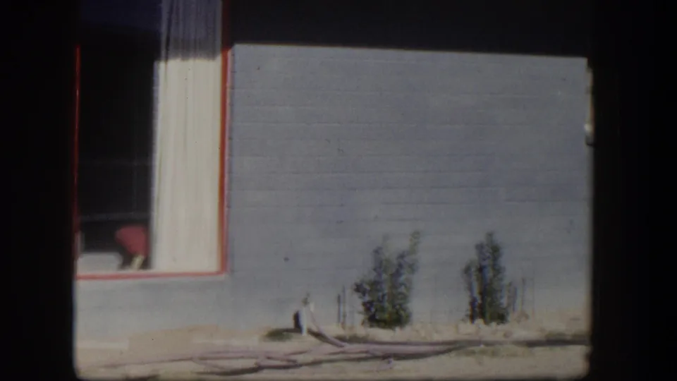1954 Nevada 8mm frame showing a house window with a red chair inside, blue-gray siding, small shrubs and a hose on sandy ground.
