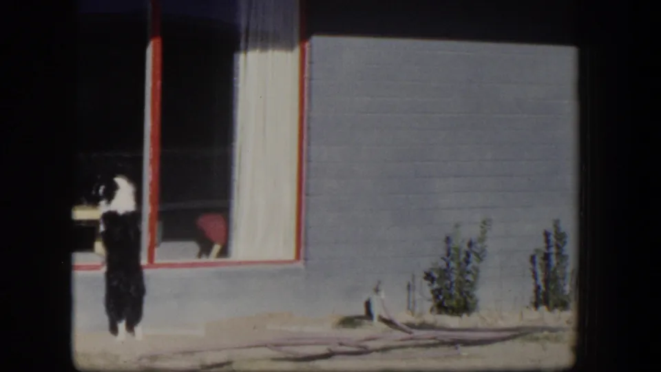 1954 Nevada home-movie frame with a person briefly visible near a doorway beside the house exterior, shrubs and garden hose.