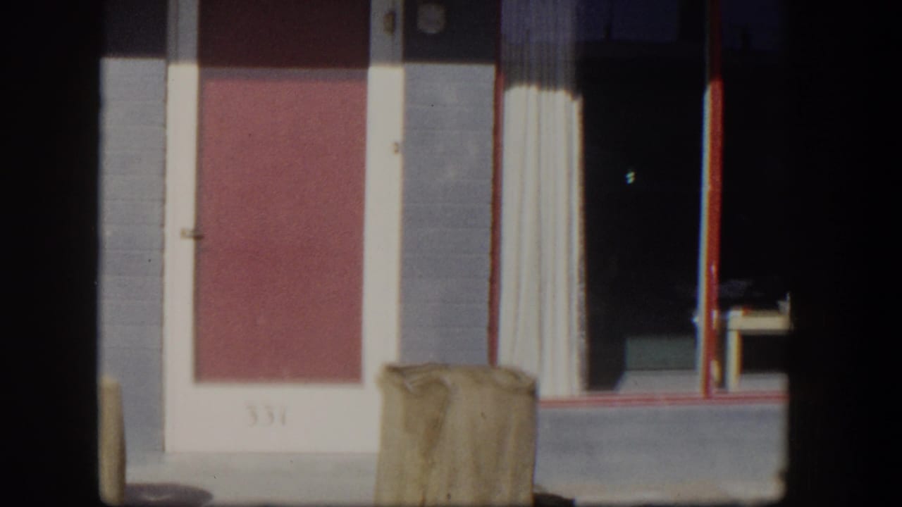 Red door, gray wall, white trim, dark window, and tan containers on a sunny porch.